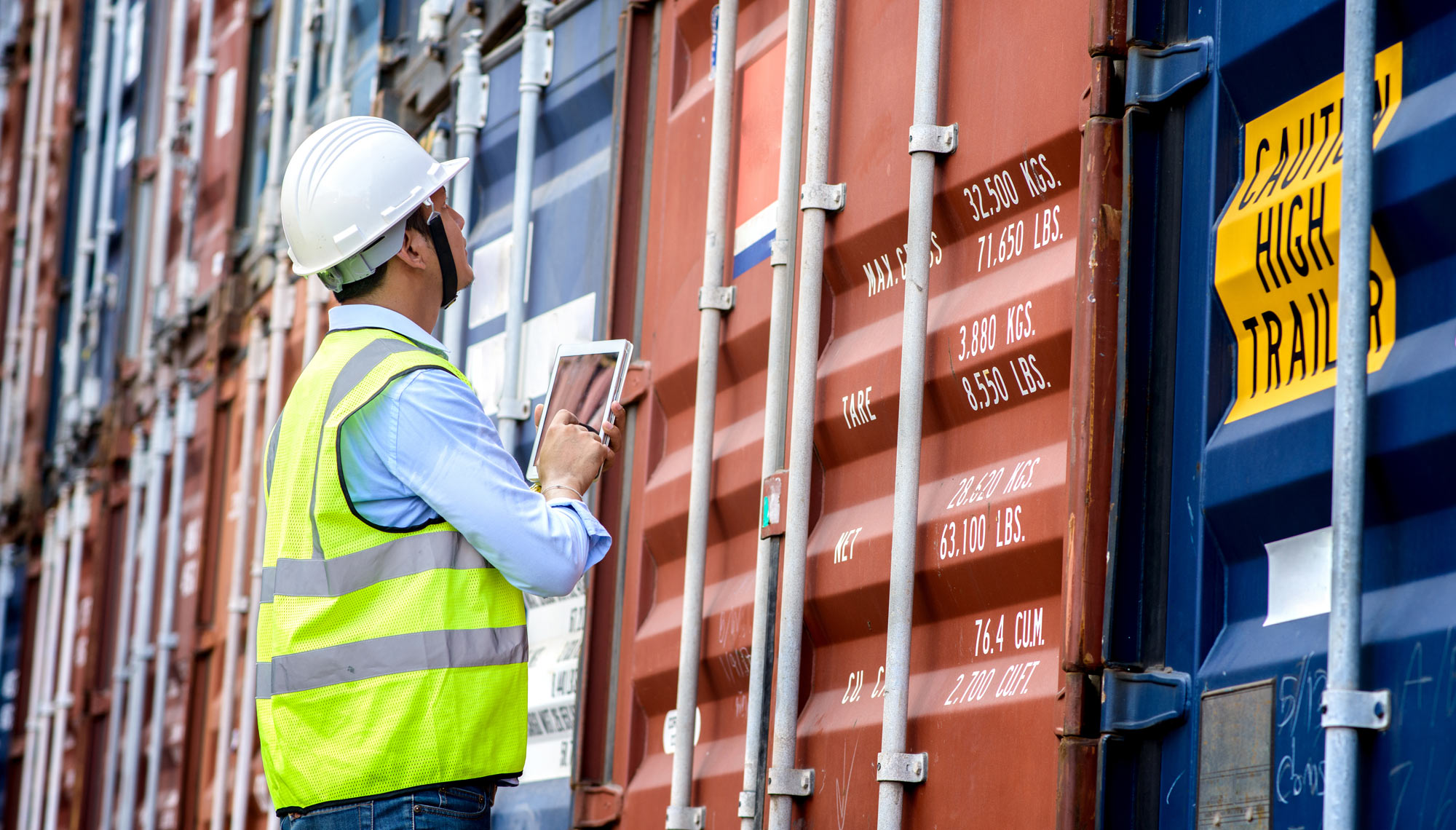 Inspector in safety gear examines shipping containers using a tablet at a port